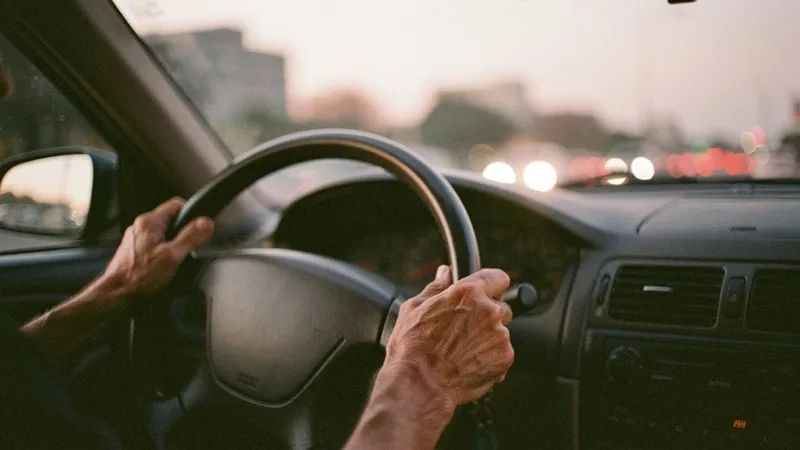 An adult driver's hands on the steering wheel at dusk, city lights blurred ahead.